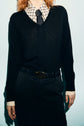 A person dressed in a black pull en laine mérinos à col V over a patterned collared shirt, black tie, and black pants with a belt. Their face is not visible.