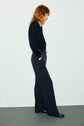 Person with long, wavy red hair stands in profile against a light background, wearing a pull en laine mérinos noir à col V, black wide-leg pants, and shiny black shoes, with hands in pockets and looking downward.