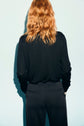A person with wavy, shoulder-length red hair is standing with their back facing the camera, wearing a black pull en laine col V and black pants against a light blue background.