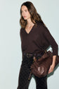 A woman with wavy brown hair wears a pull ample cachemire femme in a dark V-neck style, paired with patterned dark pants, and holds a large brown leather bag over her arm. The background is light and plain.