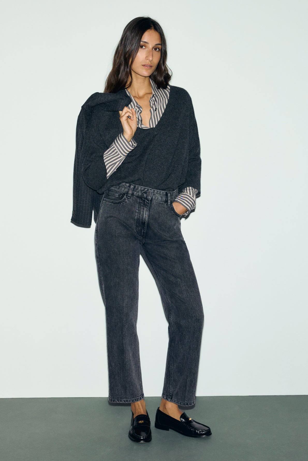 Woman stands against a pale wall, wearing a striped shirt with manches chauve-souris and a dark pull cachemire draped over her shoulders. She pairs it with gray jeans and black loafers. Her long, wavy hair frames her face as she slips one hand in her pocket.