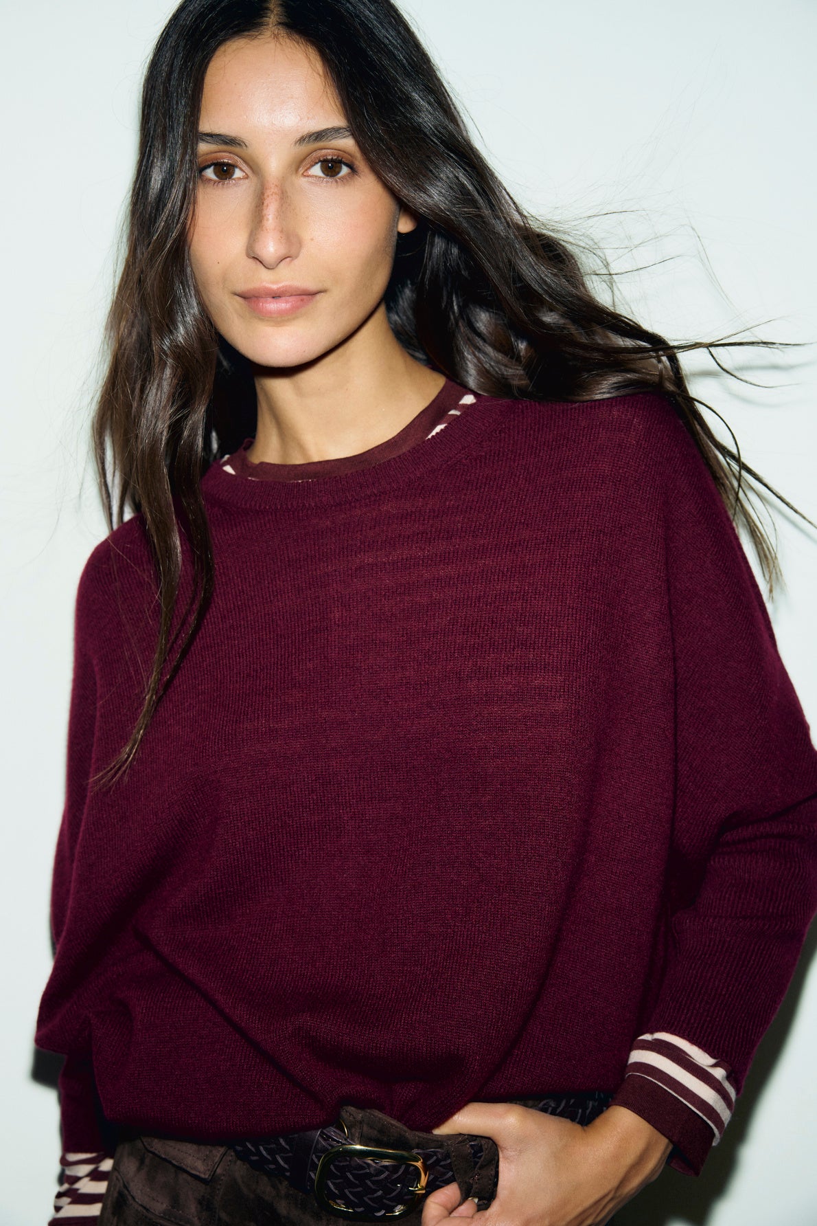 Woman with long dark hair wearing a maroon pull en cachemire with coupe ample and striped cuffs and collar, standing against a plain light background, looking at the camera with a neutral expression.