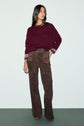 A woman stands against a plain background wearing a pull en cachemire bordeaux à manches trois-quarts, rehaussé de poignets rayés, et un pantalon en daim marron, accessoirisé d’une ceinture noire.