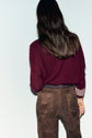 A person with long, dark hair stands facing away. They wear a maroon pull en cachemire à coupe ample with striped cuffs and high-waisted brown suede pants with a braided belt, against a plain, pale background.