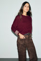 A woman wearing a burgundy pull en cachemire with striped manches trois-quarts and brown suede pants stands against a light gray background, looking confidently at the camera.