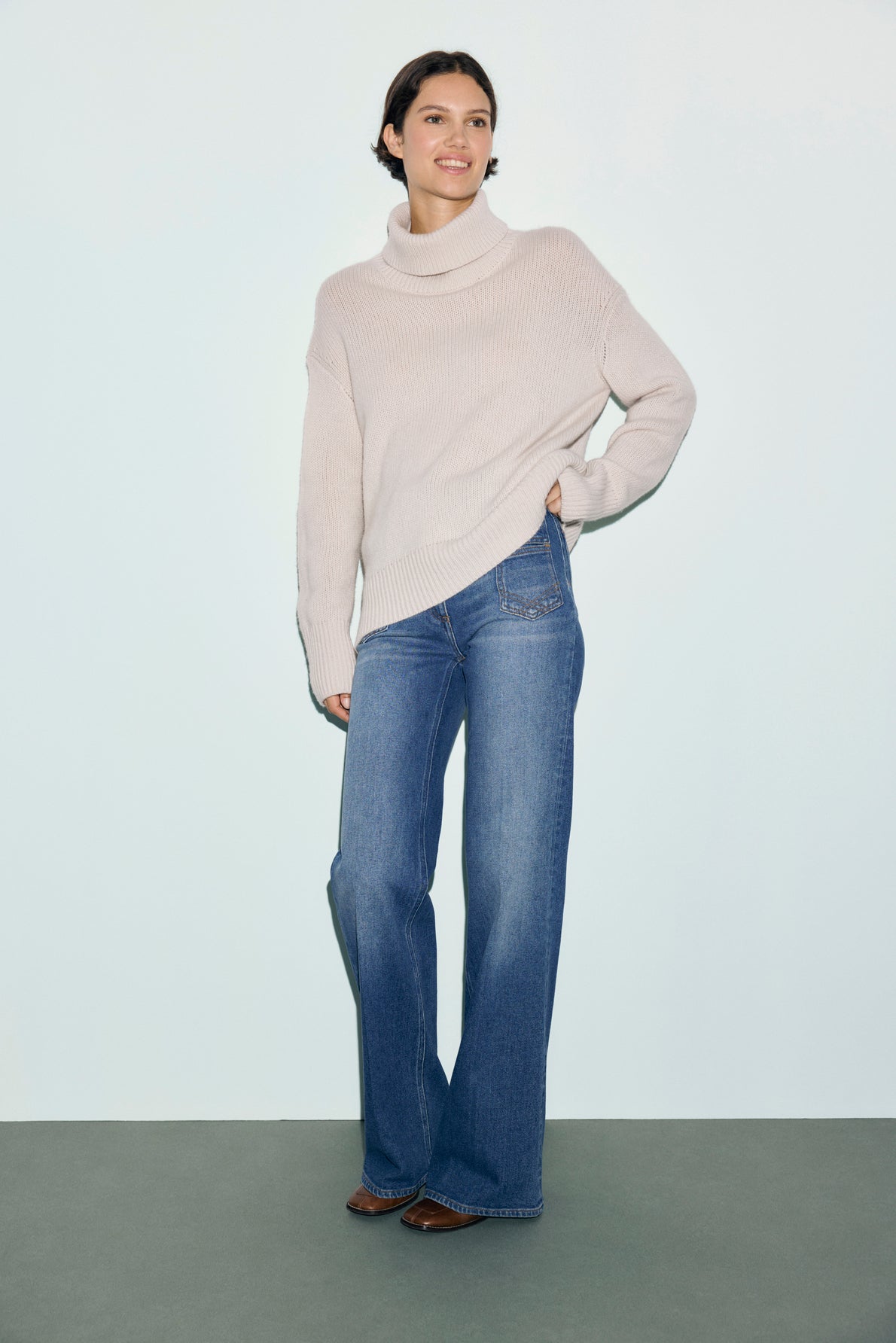 A woman stands against a plain light background, wearing a light beige col roulé sweater, blue flared jeans, and brown shoes. She is smiling with one hand on her hip.