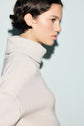 A person with short dark hair, wearing a thick, light-colored col roulé sweater, is shown in profile against a plain light background.