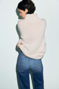 A person with short dark hair faces away, wearing a pull cachemire col roulé cream sweater and blue jeans, standing against a light background.