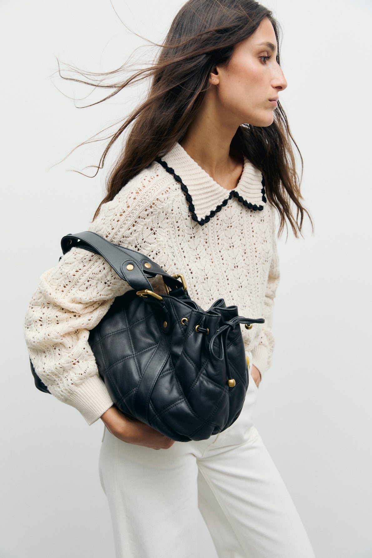 Woman in a cream pull maille crochet sweater with a black trimmed col polo and white pants, carrying a large black quilted handbag over her shoulder, stands against a plain light background.