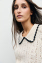 A person with long brown hair wears a cream pull maille crochet featuring a large ribbed collar trimmed in black. The background is plain and light-colored.
