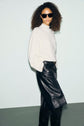 Woman with curly hair wears black sunglasses, a white pull en cachemire à col montant, and a black leather skirt. She stands against a plain light background, facing slightly to the side with hands in her pockets.