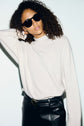 Woman with curly hair wears black sunglasses, a pull en cachemire crème à col montant and a black leather skirt with a belt. She stands against a white background with one hand behind her head.