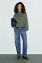 Woman with curly hair stands against a plain background, wearing an olive green pull en cachemire à col montant, blue jeans with large pockets and a coupe ample, a belt, dark loafers, and holding a black bag.