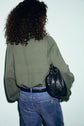 A person with curly hair, wearing a coupe ample green sweater with a col montant and blue jeans, is standing with their back to the camera, holding a black leather bag over one shoulder against a light background.