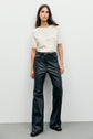 Woman standing against a plain white wall, wearing a short-sleeved white lace top with un délicat motif ajouré and high-waisted black faux leather flared pants with black shoes.