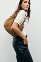 A woman stands sideways, wearing a white short-sleeve pull en coton au motif ajouré, black leather pants, and carrying a large tan suede shoulder bag. She has long brown hair and her hand is in her pocket. The background is plain white.