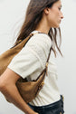 A woman with long brown hair wears a cream pull en coton à maille point de riz and black pants, carrying a large tan suede bag over her shoulder. She is facing sideways against a plain light background.