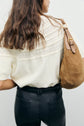 A woman with wavy brown hair wears a cream lace top reminiscent of maille point de riz, black high-waisted pants, and carries a brown suede handbag over her shoulder, facing away from the camera.