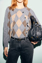 Woman wearing a gray and beige argyle sweater with black jeans holds a black woven leather handbag, standing against a pale blue background. The motif losanges of her gilet laine adds a touch of classic elegance to the look.