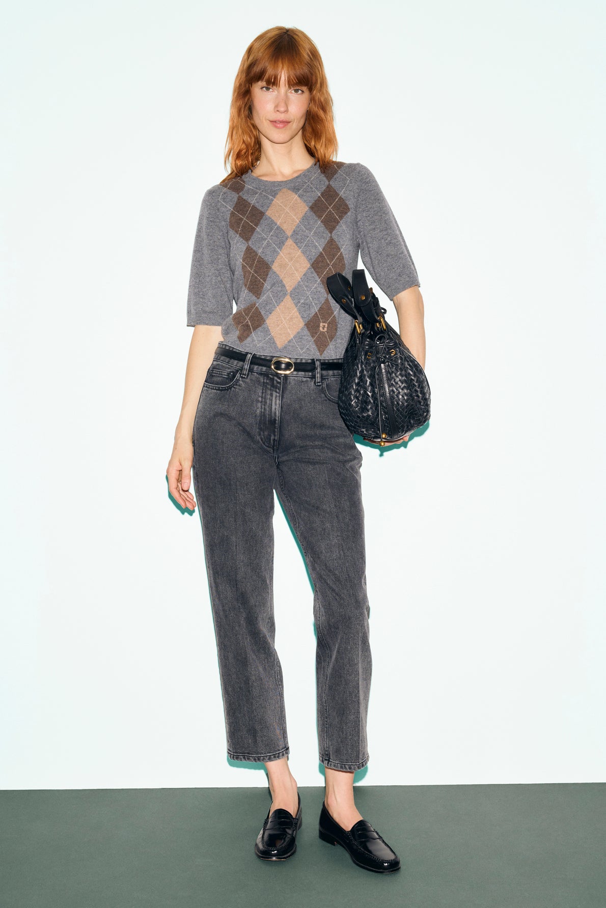 A woman with red hair wears a gray pull laine à losanges, high-waisted gray jeans, and black loafers. She holds a textured black bag in her right hand and stands against a plain light background.