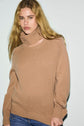 A person with long, wavy blonde hair wears a pull en laine camel à manches longues featuring a high collar and blue jeans, standing against a plain, light background.