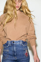 A woman with long, wavy blonde hair obscuring part of her face, wearing a pull en laine camel à manches longues and blue denim jeans featuring a patch with an Eiffel Tower illustration.