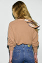 A person with long, wavy blonde hair wearing a pull en laine camel à manches longues, blue jeans, and a patterned col amovible scarf stands facing away from the camera against a plain white background.