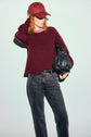 Person in a pull bordeaux en laine and matching cap poses against a light background, holding a black textured bag and wearing black jeans with a belt. Their other hand is touching the back of their head, while the subtle col amovible on their sweater adds an extra touch of style.