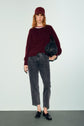 Woman in a burgundy cap and pull en laine bordeaux, gray jeans, and black loafers stands holding a black textured bag against a plain background.