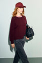 A person with red hair wearing a pull bordeaux en laine, matching burgundy cap, dark jeans, and holding a black textured handbag walks against a light background.