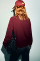 A person with wavy, reddish hair is wearing a pull en laine bordeaux and a matching baseball cap. They’re holding a black, textured bag and facing away from the camera against a plain background.