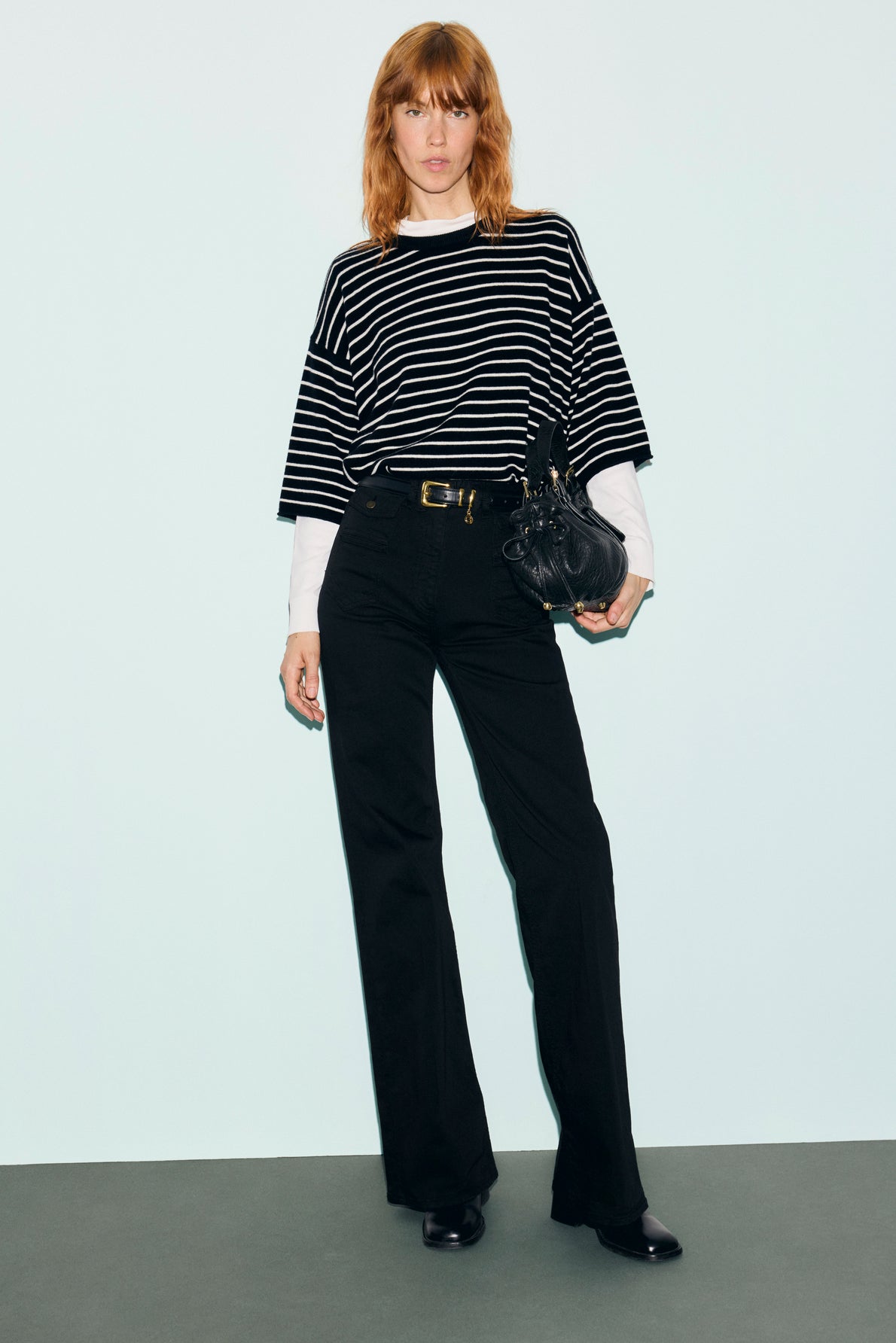 A person with light brown hair wears a pull rayé laine—a black and white striped knit sweater—over a white long sleeve, paired with black pants, black shoes, and holds a black handbag, standing against a plain light-colored wall.