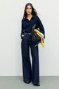 Woman with long brown hair wearing a pull bleu marine en laine mérinos with a chic col polo and wide-leg jeans, holding a black quilted bag with a yellow scarf, standing against a plain light background.