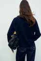A person with long brown hair, seen from behind, wears a pull bleu marine en laine mérinos and dark jeans, holding a woven black handbag with a yellow scarf tied to it. The background is plain and light-colored.