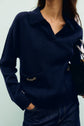 A person wearing a navy blue knit sweater crafted from laine mérinos, featuring a classic col polo and two front pockets, each adorned with a small gold chain, paired with dark jeans and holding a black handbag.