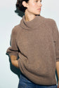 Person wearing a cozy brown col roulé ouvert sweater in soft laine and blue jeans, looking to the side with one hand casually placed in a pocket, against a light background.