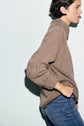 Person with short dark hair wearing a brown pull en laine and blue jeans stands sideways against a light background, with one hand in their pocket.