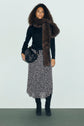 Woman stands against a plain background wearing a pull col roulé noir in laine mérinos, a long patterned skirt, black boots, a large brown scarf, and holding a black bag.