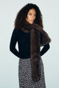 A woman with curly hair wears a pull noir en laine mérinos, a long dark brown faux fur scarf, and a black skirt with a white polka dot pattern, standing against a plain light background.