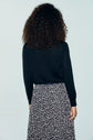 A person with curly hair is standing facing away, wearing a long-sleeve pull noir in laine mérinos and a black-and-white patterned skirt against a plain light background.