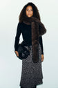 A woman wearing a pull col roulé noir en laine mérinos, a long polka dot skirt, a large brown faux fur scarf, and carrying a black bag stands against a plain light background.
