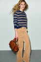 A person with long wavy hair wears a navy striped motif rayé sweater in laine ultra douce and beige corduroy pants, carrying a brown leather handbag, standing against a plain light background.
