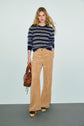 A woman stands against a plain background wearing a motif rayé pull Rima in navy and beige, paired with tan wide-leg corduroy pants and platform shoes. She carries a brown handbag and has long blonde hair, while the laine ultra douce of her sweater adds an extra touch of comfort to her look.