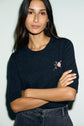 Woman with long dark hair wears a pull en laine à col rond, featuring a small embroidered motif fleuri on the chest, standing with arms crossed against a light background.