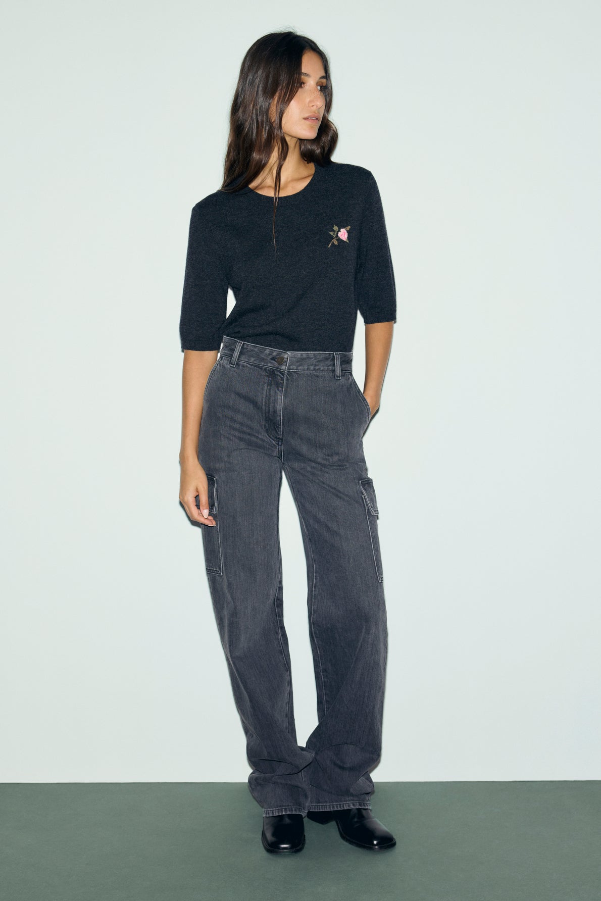 A woman stands against a light background wearing a dark short-sleeve pull en laine à col rond with a small motif fleuri embroidered design, loose-fitting gray cargo pants, and black shoes.