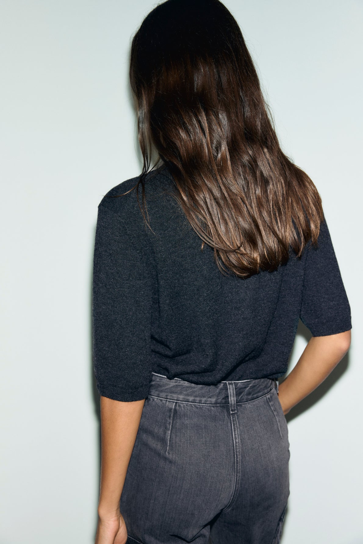 A person with long brown hair, wearing a dark short-sleeve top with un col rond and high-waisted gray jeans, stands facing away from the camera against a plain light background.
