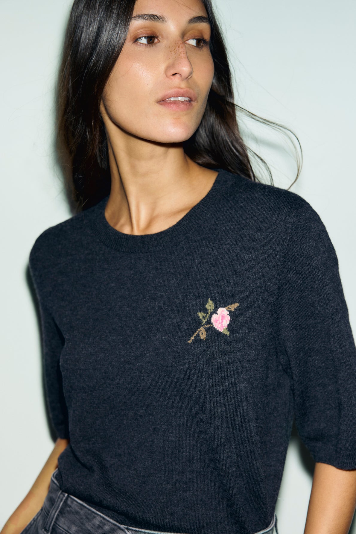 Woman with long dark hair wears a pull en laine with a col rond and a small, embroidered pink rose motif fleuri on the chest, standing against a pale background.