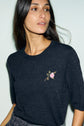 Woman with long dark hair wears a pull en laine with a col rond and a small, embroidered pink rose motif fleuri on the chest, standing against a pale background.