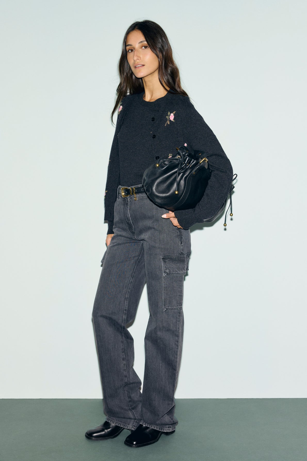 Woman stands against a light background wearing a dark long-sleeve top with motif fleuri embroidery, wide-leg cargo jeans, black boots, and holding a black bag. She has long, wavy dark hair and a relaxed expression.