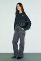 Woman stands against a light background wearing a dark long-sleeve top with motif fleuri embroidery, wide-leg cargo jeans, black boots, and holding a black bag. She has long, wavy dark hair and a relaxed expression.
